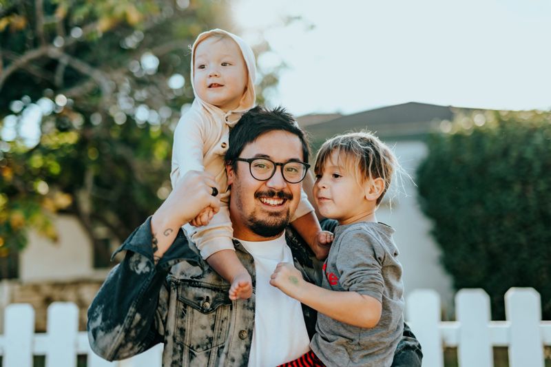 Vater mit 2 kleinen Kindern auf Arm und Schultern vor Wohnhaus